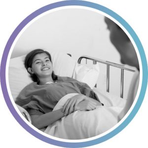 A young woman with dark hair lays in a hospital bed, smiling at the healthcare worker next to her bed.