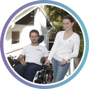 A young male wheelchair user and his female partner are outside their home on the accessible ramp smiling at camera.