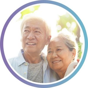 An older couple with their arms around each other in a sunlit park, looking into the medium distance.