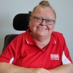 Head and shoulders photo of woman smiling at camera, she wears glasses and a red t-shirt and her hair is in a bun.