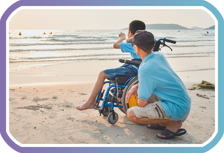 A young boy wheelchair user is on the shoreline of a beach with his adult male career, they are both looking out towards the people swimming as the young boy points to them.