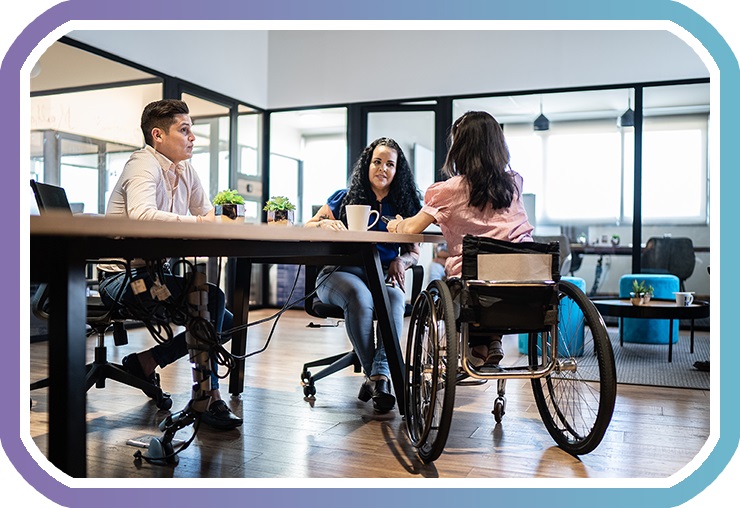 A male and two females, one a wheelchair user, talk around a meeting table in a well-lit office space.